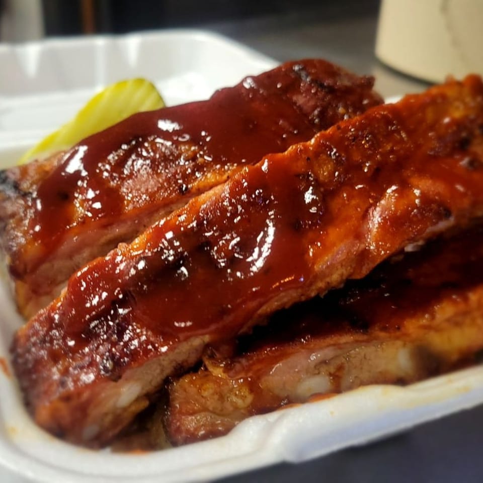 Stuboy's BBQ ribs, glistening with sauce, served in a white takeout container with a pickle.