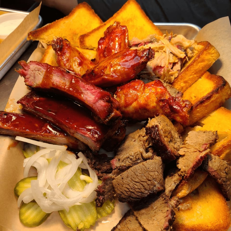 A platter of Stuboy's BBQ with ribs, chicken wings, brisket, pulled pork, Texas toast, pickles, and onions.