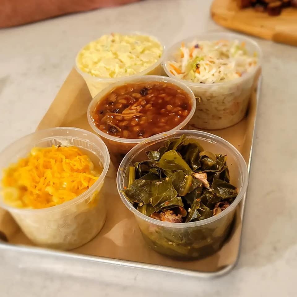 Five plastic containers of Stuboy's BBQ sides: mac and cheese, baked beans, collard greens, potato salad, and coleslaw.